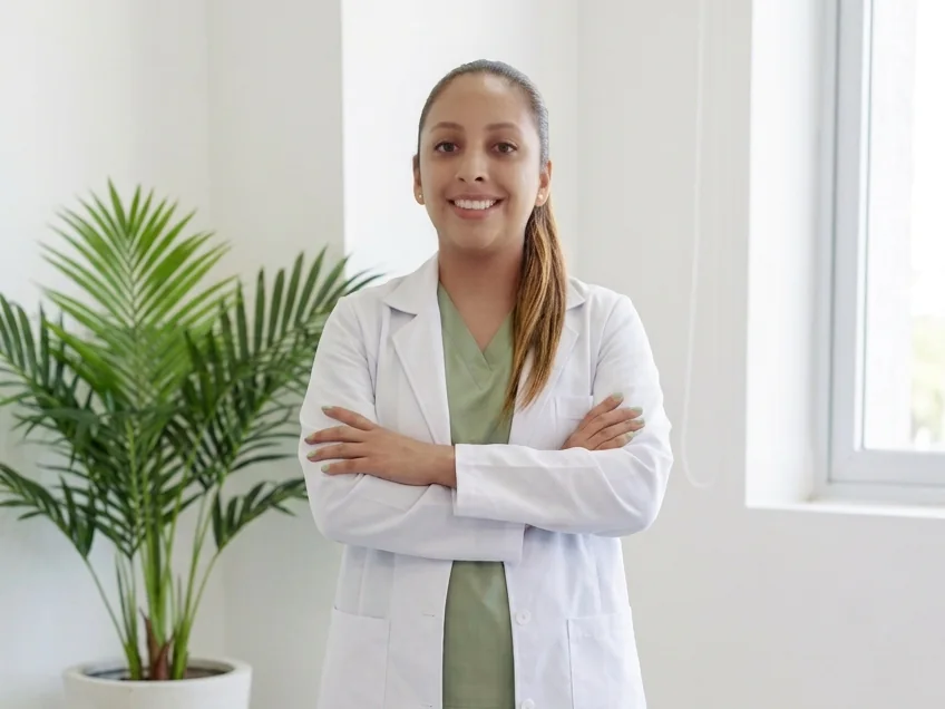 Paola Beltrán Méndez con bata blanca, de pie y con los brazos cruzados en su consultorio