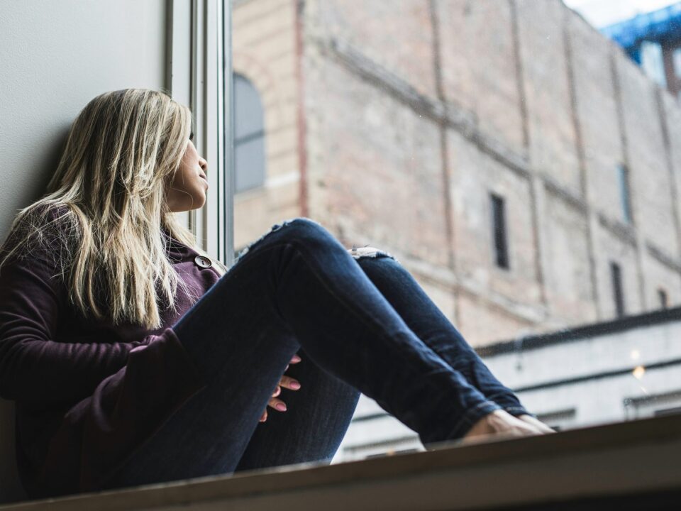 Mujer sentada junto a una ventana, mirando hacia afuera en actitud pensativa